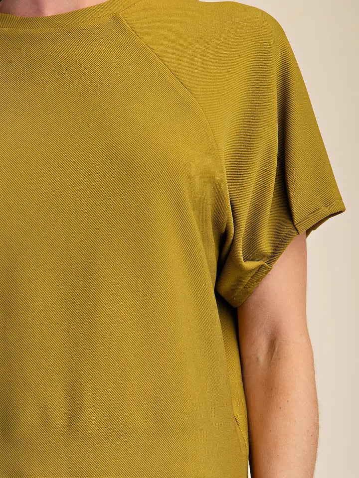 Close-up of a person wearing a short-sleeve textured knit top in a mustard yellow color, showing the shoulder seam and sleeve detail against a plain beige background.