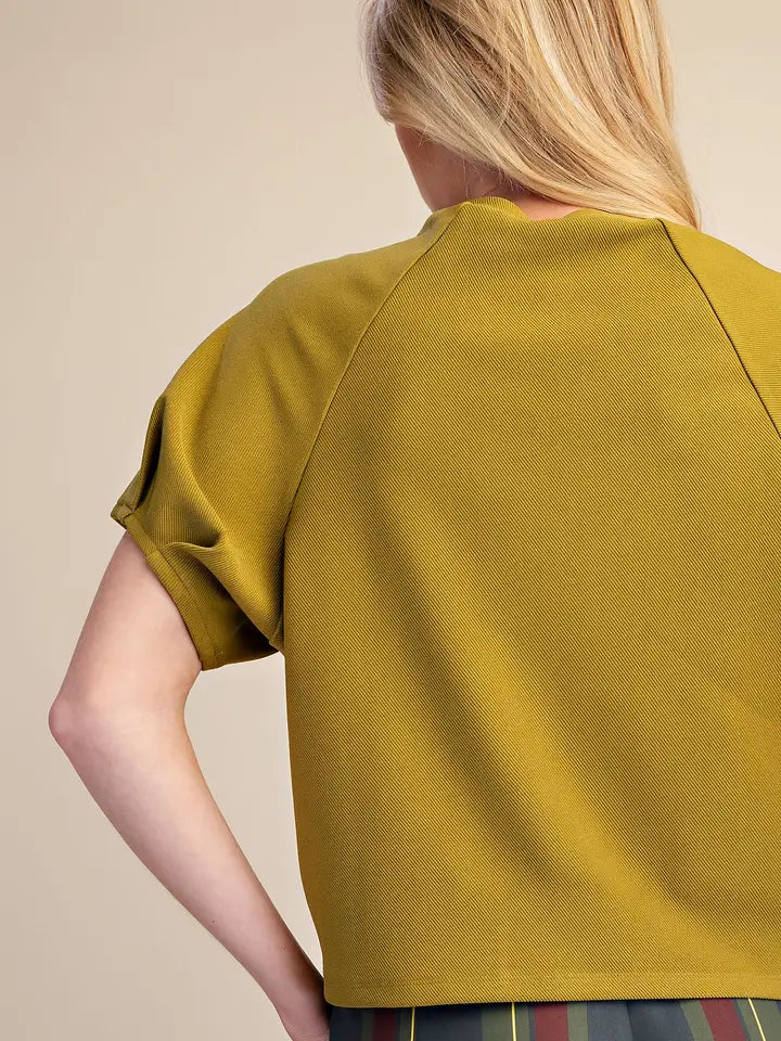 A woman shown from the back wearing a mustard yellow short-sleeve top, photographed against a plain beige studio background.