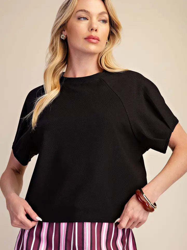 A blonde woman wearing a black short-sleeve top with raglan sleeves and a striped skirt, posing against a solid beige background.