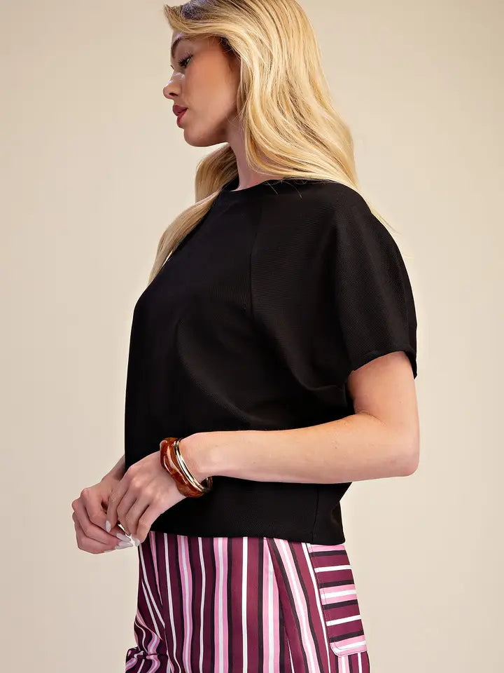 A woman shown in side profile wearing a black short-sleeve top and pink, purple, and white vertically striped trousers, accessorized with stacked gold and brown bracelets, against a plain beige studio background.