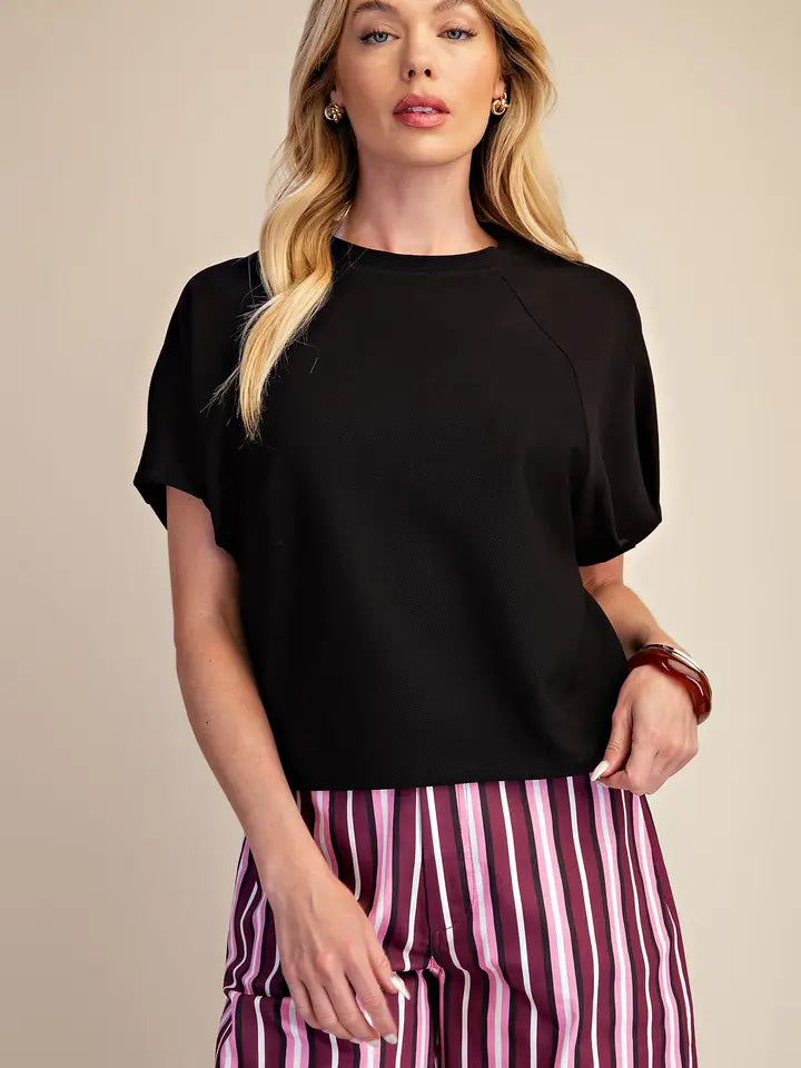 A woman wearing a black short-sleeve crewneck top and pink, purple, and white vertically striped pants, standing against a solid beige background.