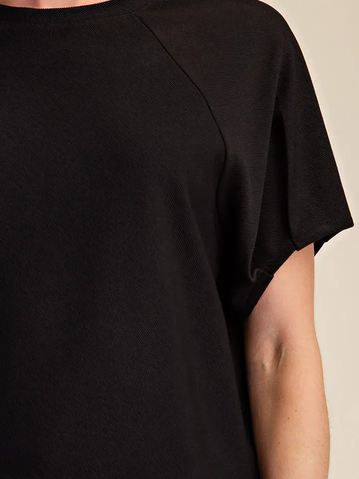 Close-up of a person wearing a black short-sleeve top, showing the shoulder seam and part of the arm against a plain light beige background.
