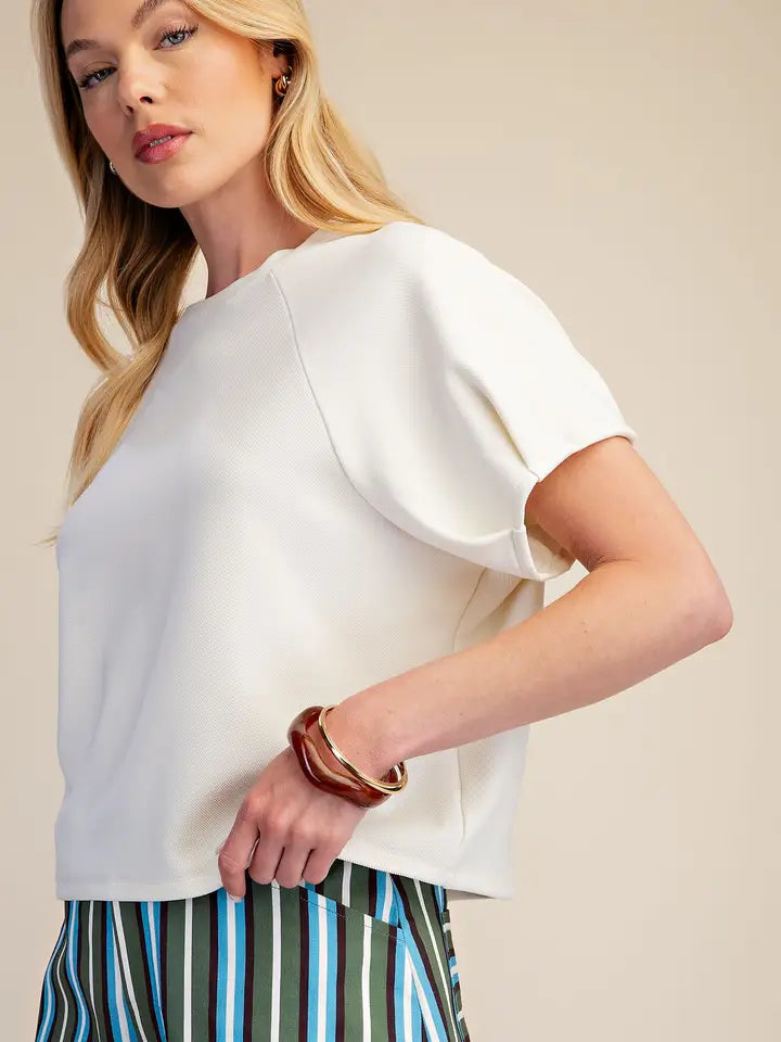 A blonde woman wearing a cream-colored short-sleeve top with balloon sleeves and striped trousers, accessorized with a brown leather bangle, against a solid beige background.