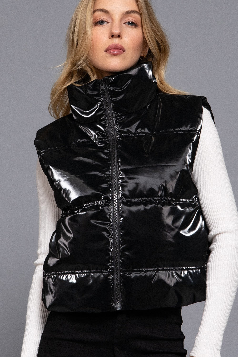 A woman wearing a shiny black puffer vest over a white long-sleeve top, photographed against a plain gray studio background.