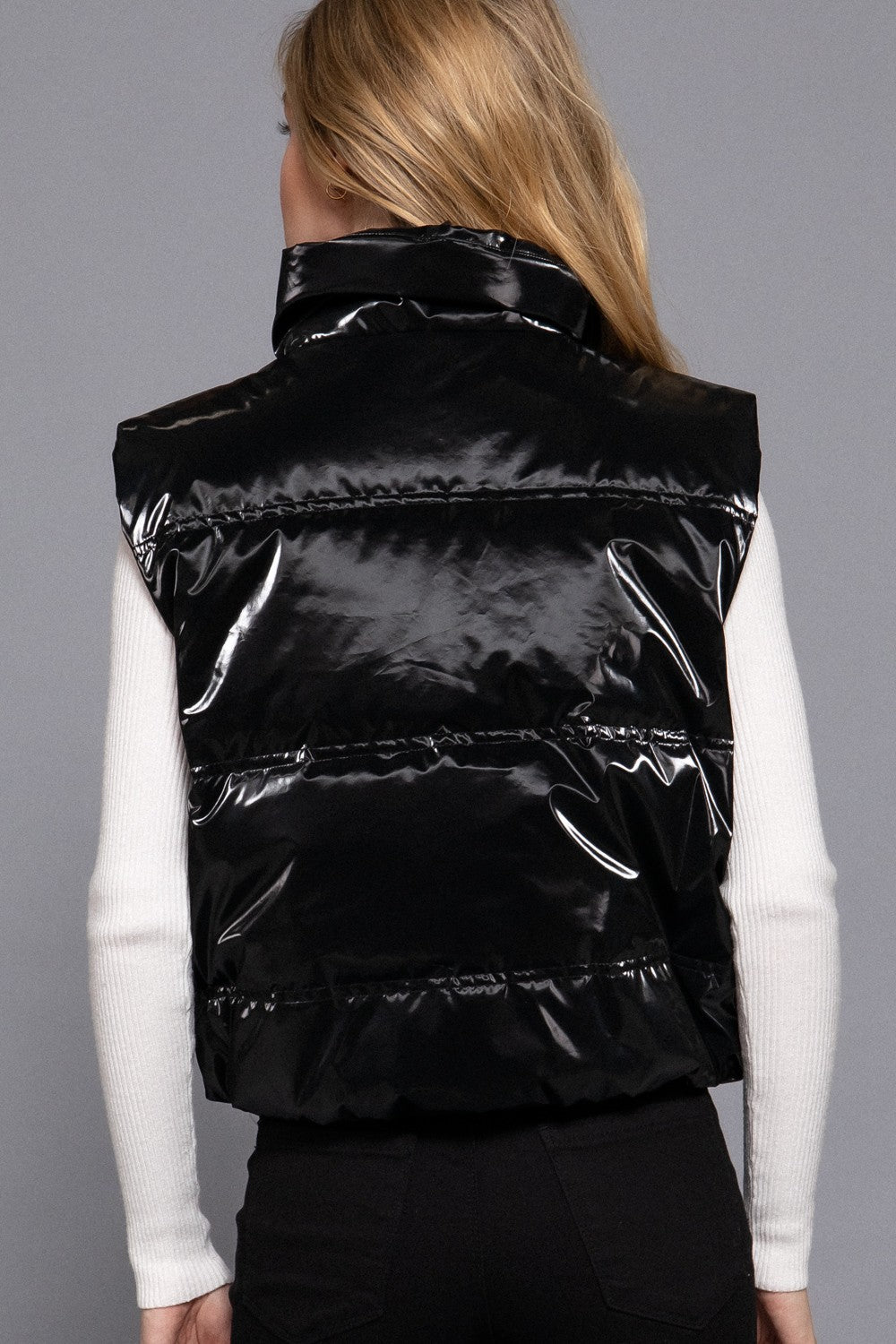 A woman viewed from the back wearing a shiny black puffer vest over a white long-sleeve top and black pants, against a plain gray studio background.
