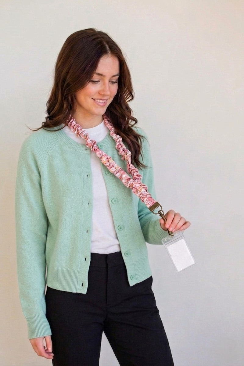 A woman wearing a mint green cardigan, white t-shirt, and black pants models a pink and white floral patterned fabric lanyard with a clear ID badge holder.