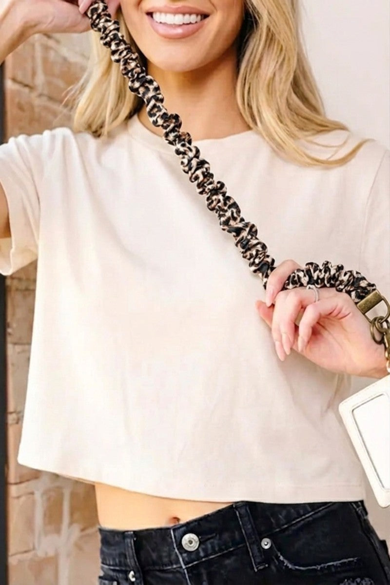 A woman wearing a cream-colored short-sleeve crop top and dark denim jeans, holding a leopard-print chain strap across her body.