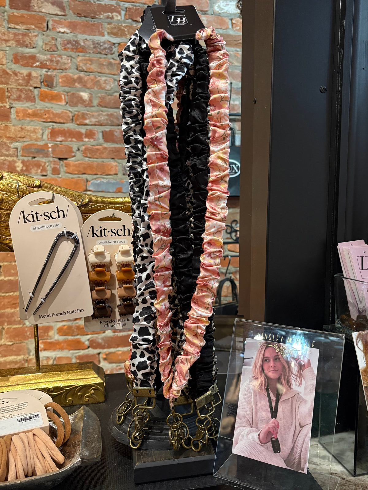 A display of three ruffled  lanyards hanging on a black stand in a retail setting. The products feature three distinct patterns: a black and white leopard print, a solid black with gold chain accents, and a pink and white leopard print.