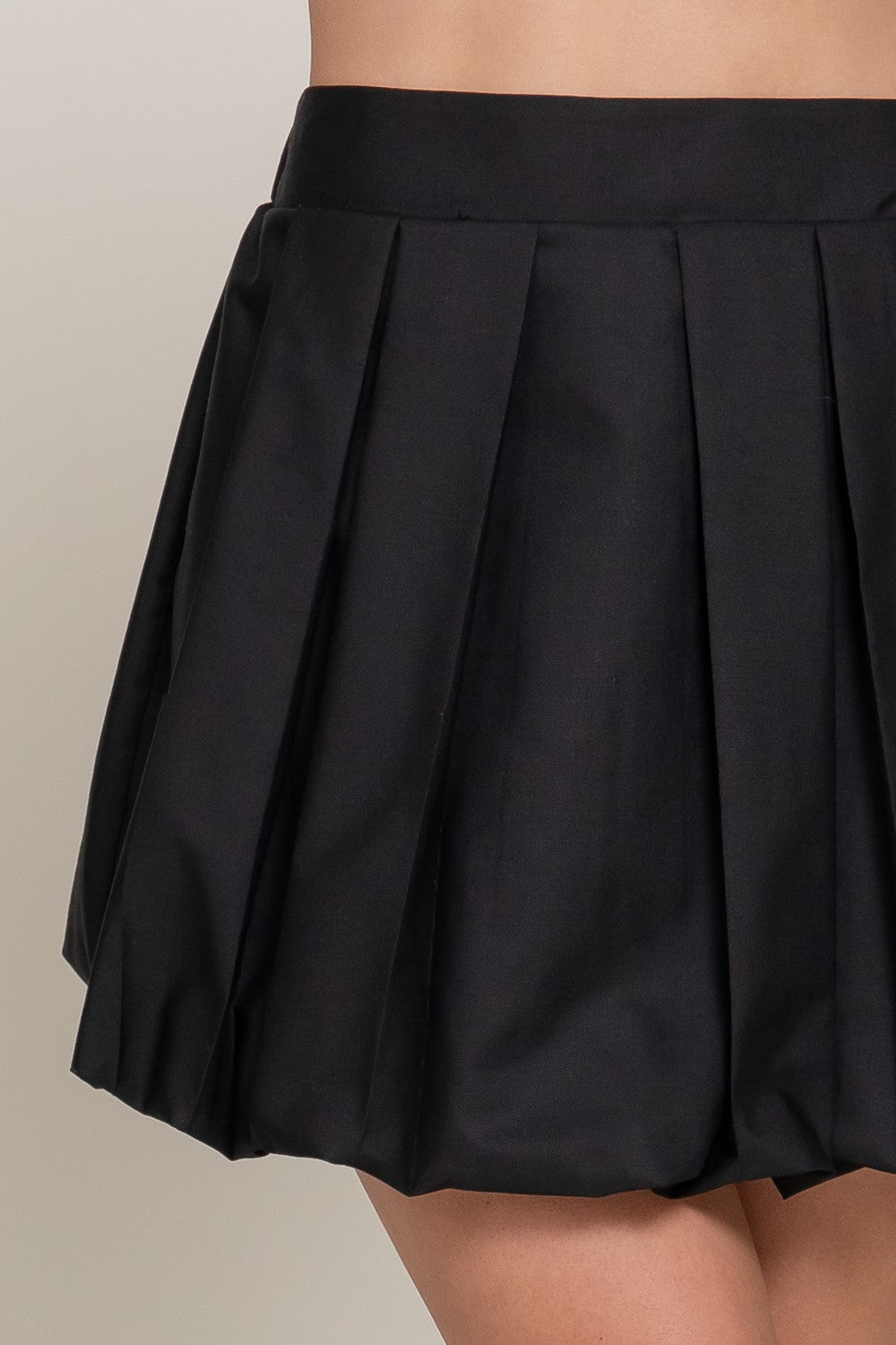 Close-up of a black pleated mini skirt worn by a model, showing the fabric texture and pleated details against a neutral light gray background.