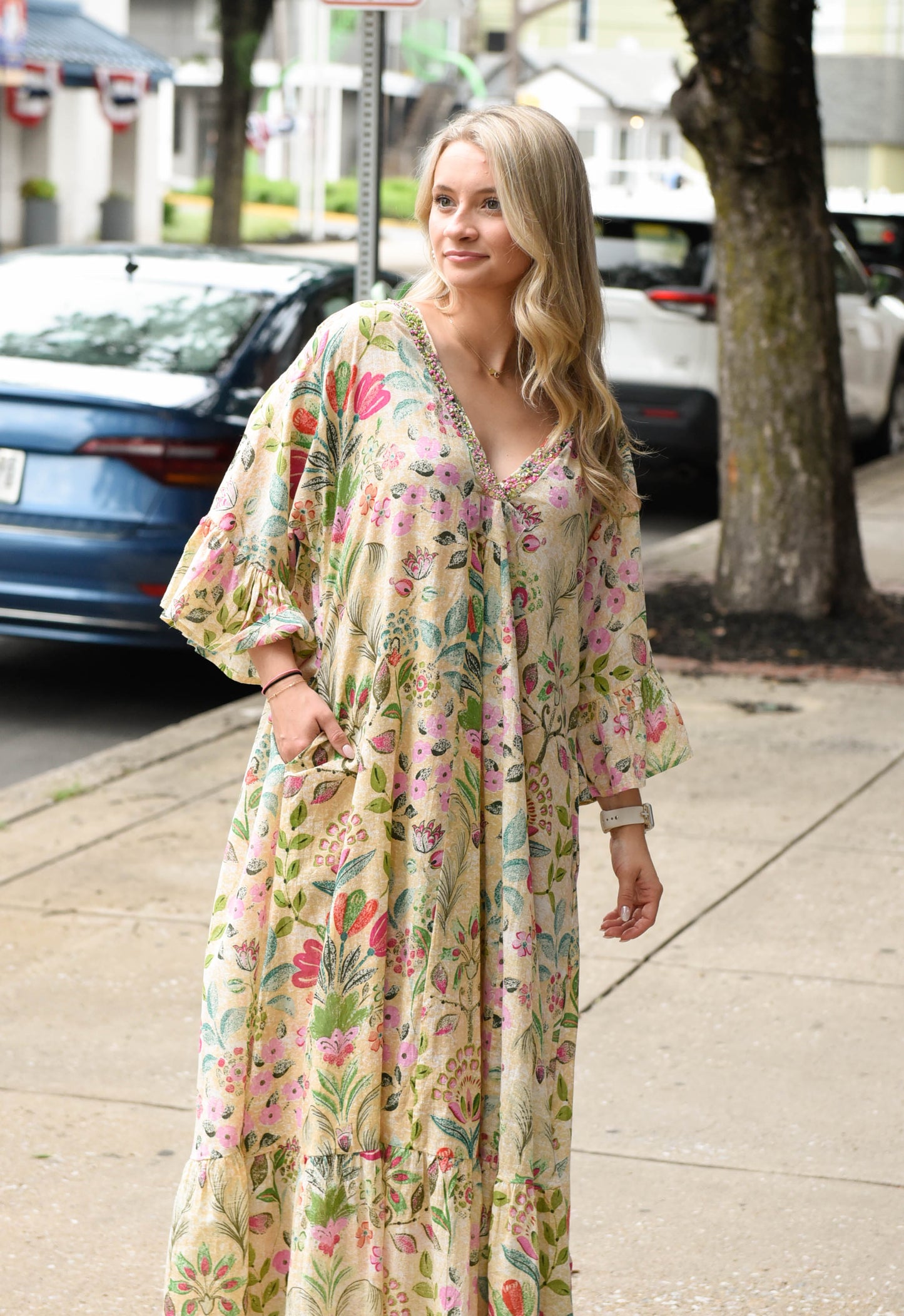 A woman wearing a long, flowy maxi dress with a colorful floral print on a cream background, standing on a city sidewalk next to a street with parked cars.