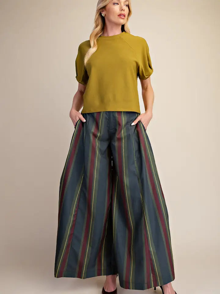 A woman modeling a mustard yellow short-sleeve top and wide-leg striped pants with vertical stripes in dark green, burgundy, and yellow, standing against a solid beige studio background.