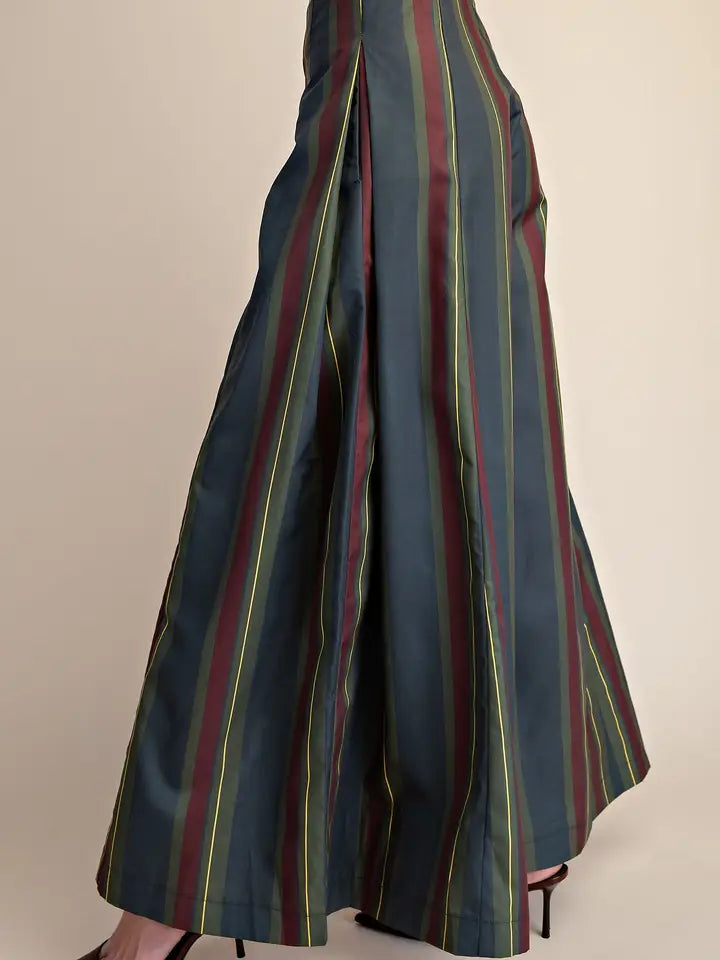 A close-up of a person wearing wide-leg pleated trousers with vertical stripes in dark teal, burgundy, olive green, and yellow, paired with black high heels against a plain beige background.