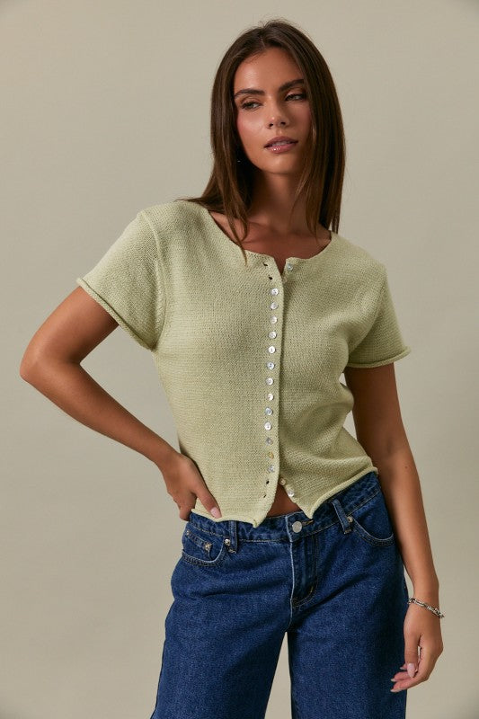 Woman wearing a green short-sleeve cardigan and blue jeans against a neutral background