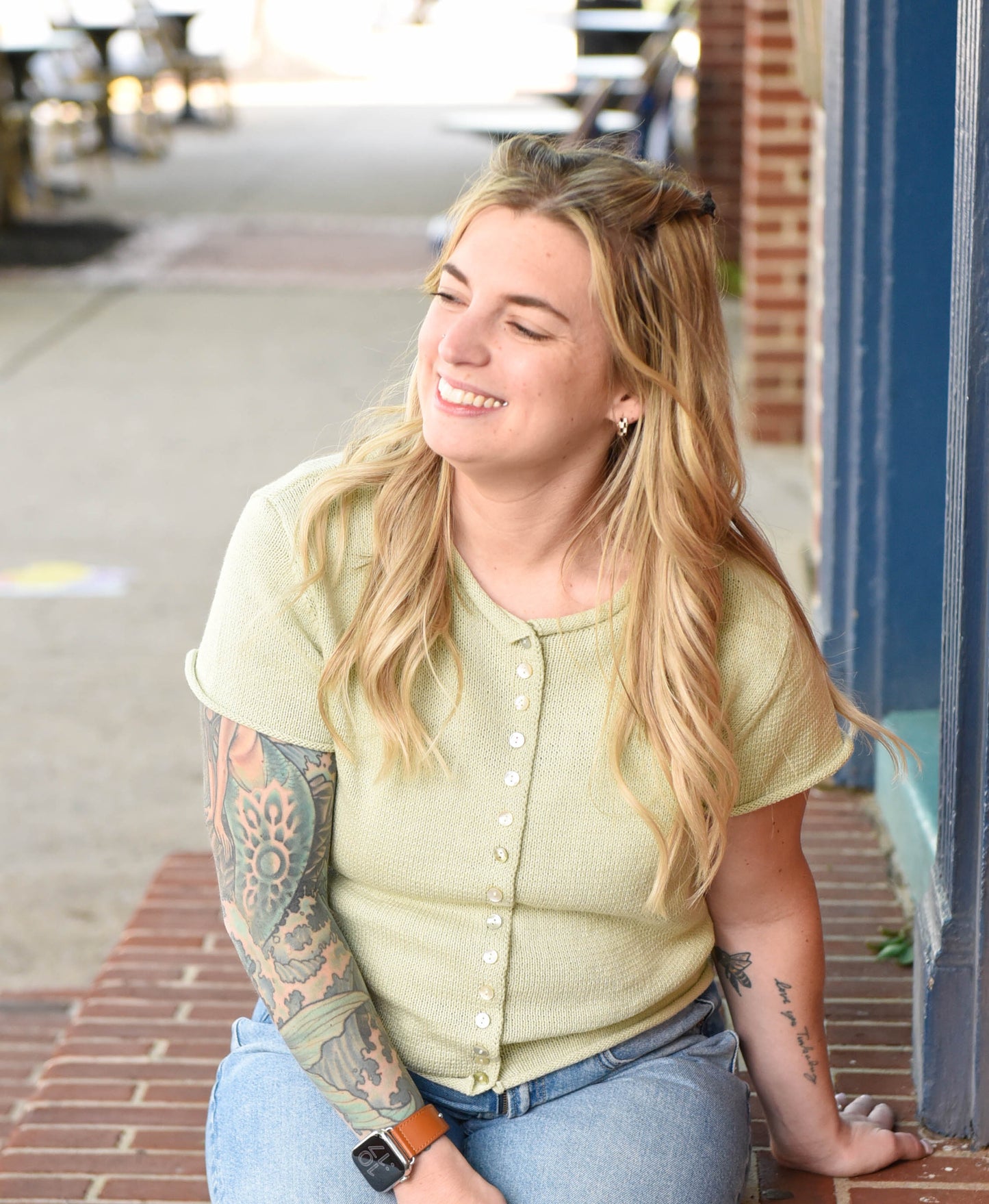 Woman with tattoos wearing a light green shirt and blue jeans sitting on a step.
