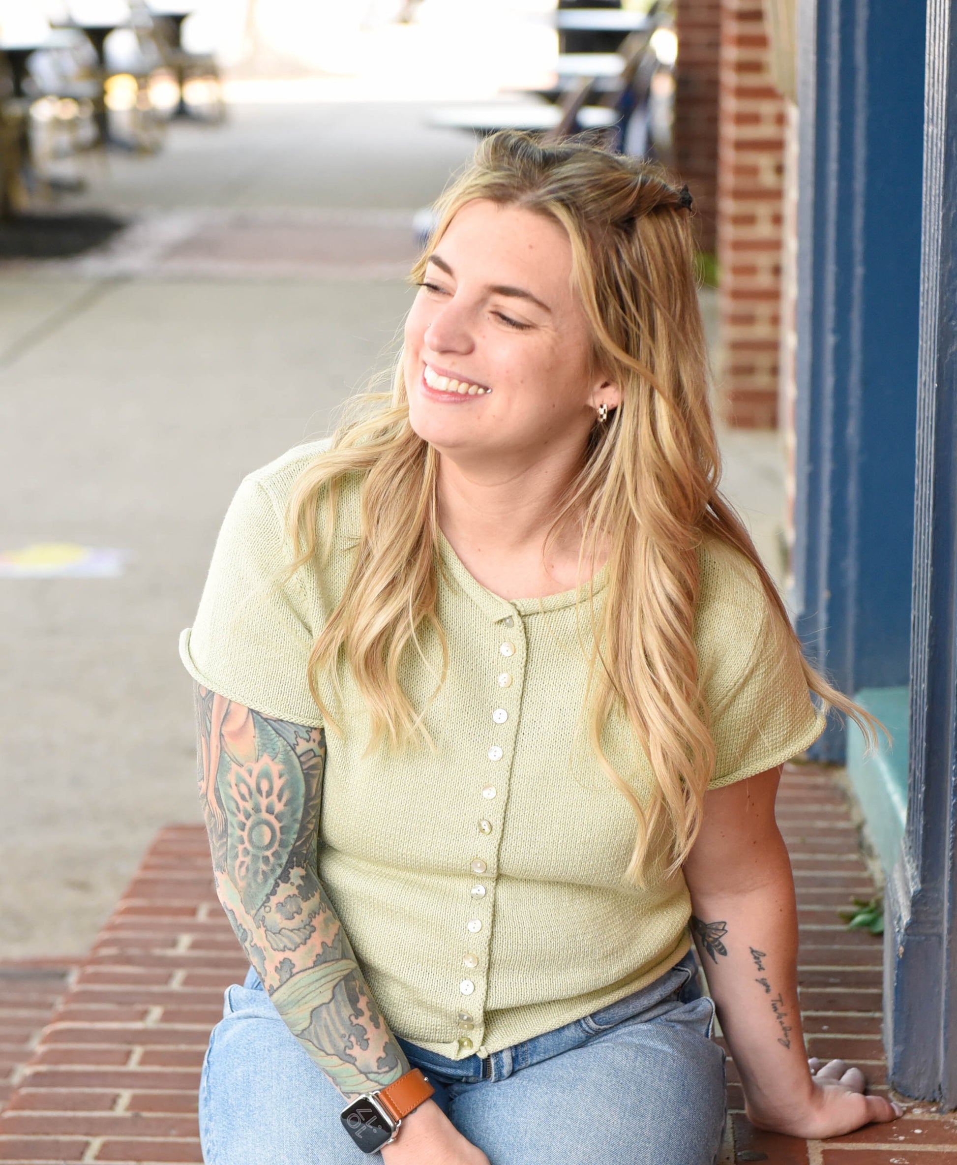 Woman with tattoos wearing a light green shirt and blue jeans sitting on a step.