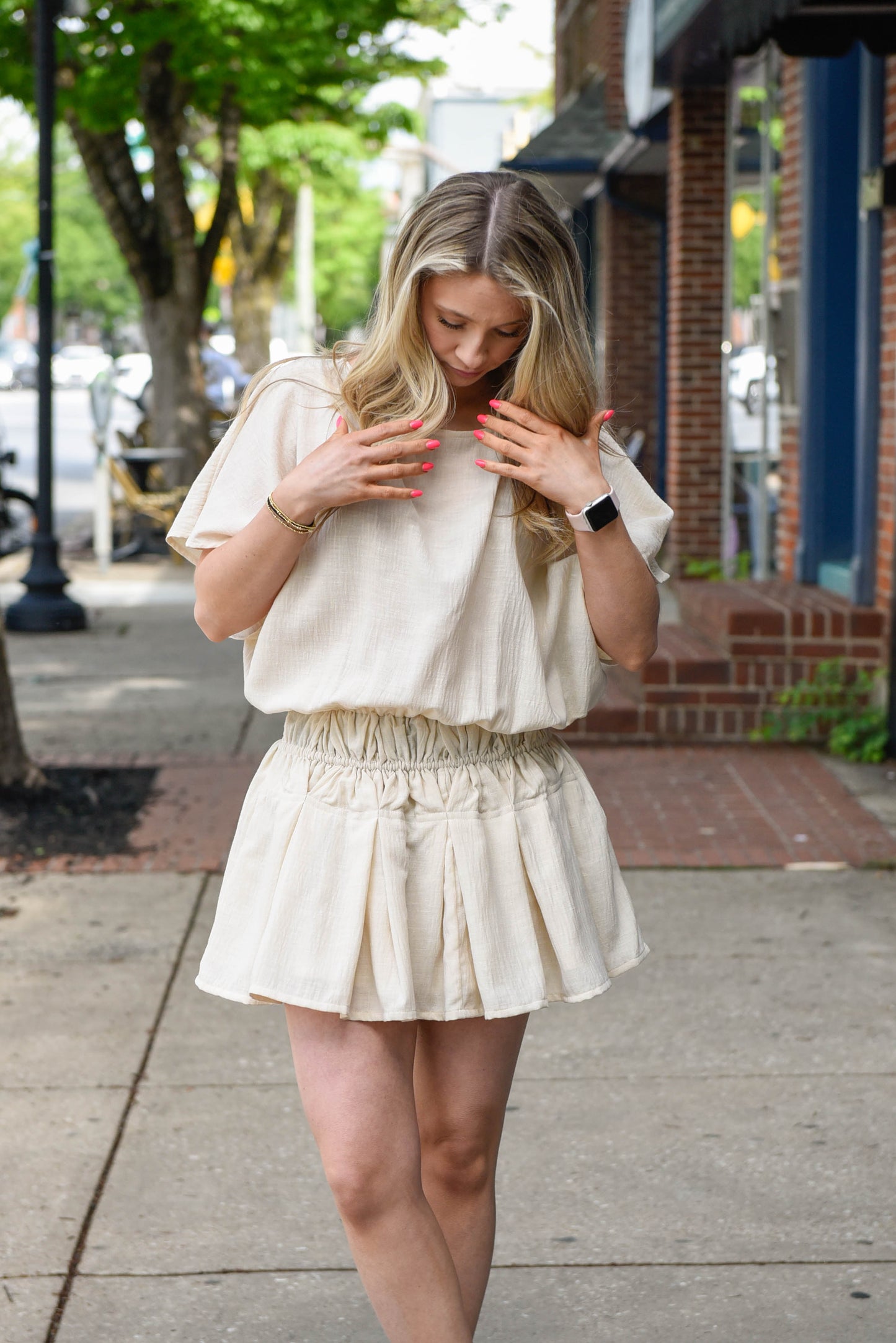 Woman in a light-colored outfit walking on a sidewalk with buildings and trees in the background