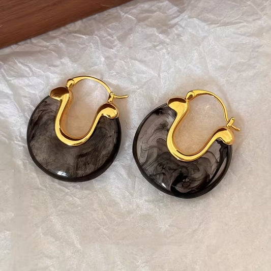 A pair of gold hoop earrings with dark gray and black marbled resin discs, displayed on a crumpled white paper background.