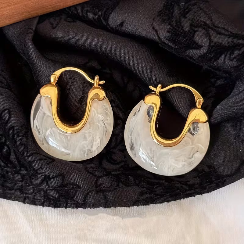A pair of hoop earrings with gold-toned metal and translucent white marbled stone discs, displayed on black patterned fabric.