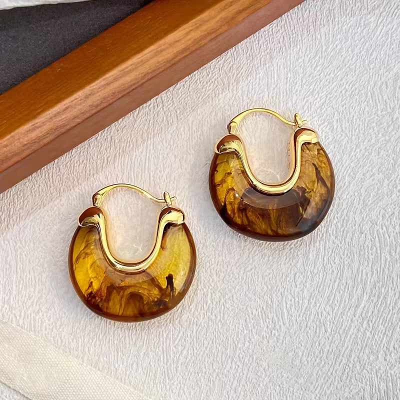 A pair of gold-tone hoop earrings with marbled amber-brown resin inlays, displayed on a textured white surface next to a wooden box edge.