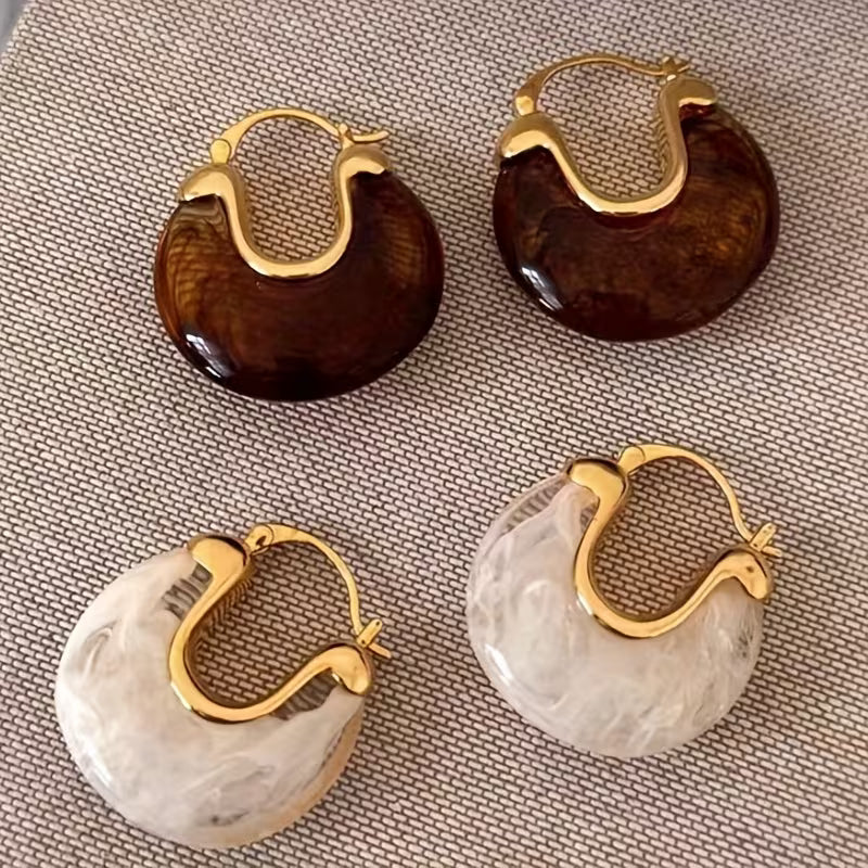 Four large hoop earrings arranged on a textured beige fabric background. Two hoops have glossy dark brown marbled resin with gold-tone metal hoops, and two have marbled off-white resin with gold-tone metal hoops.