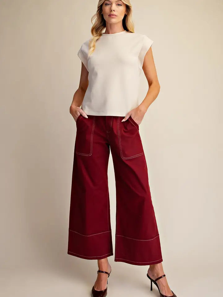 A woman modeling a white sleeveless top and deep burgundy wide-leg cropped pants with contrast stitching, standing against a plain beige studio background.