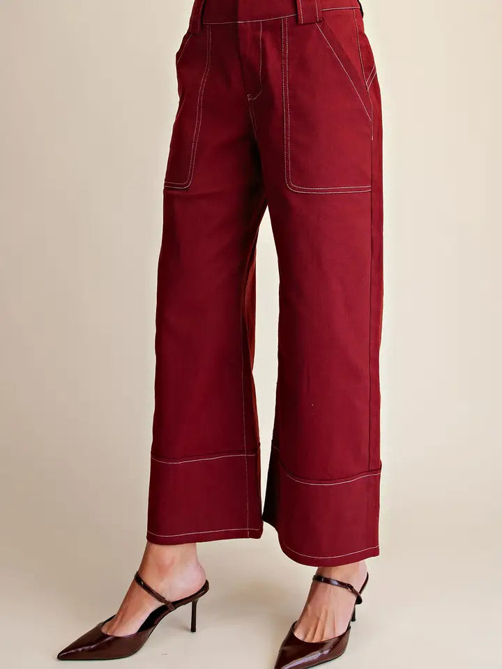 A person wearing burgundy cropped wide-leg pants with contrast white stitching and dark brown pointed-toe mule heels, shown against a plain beige studio background.