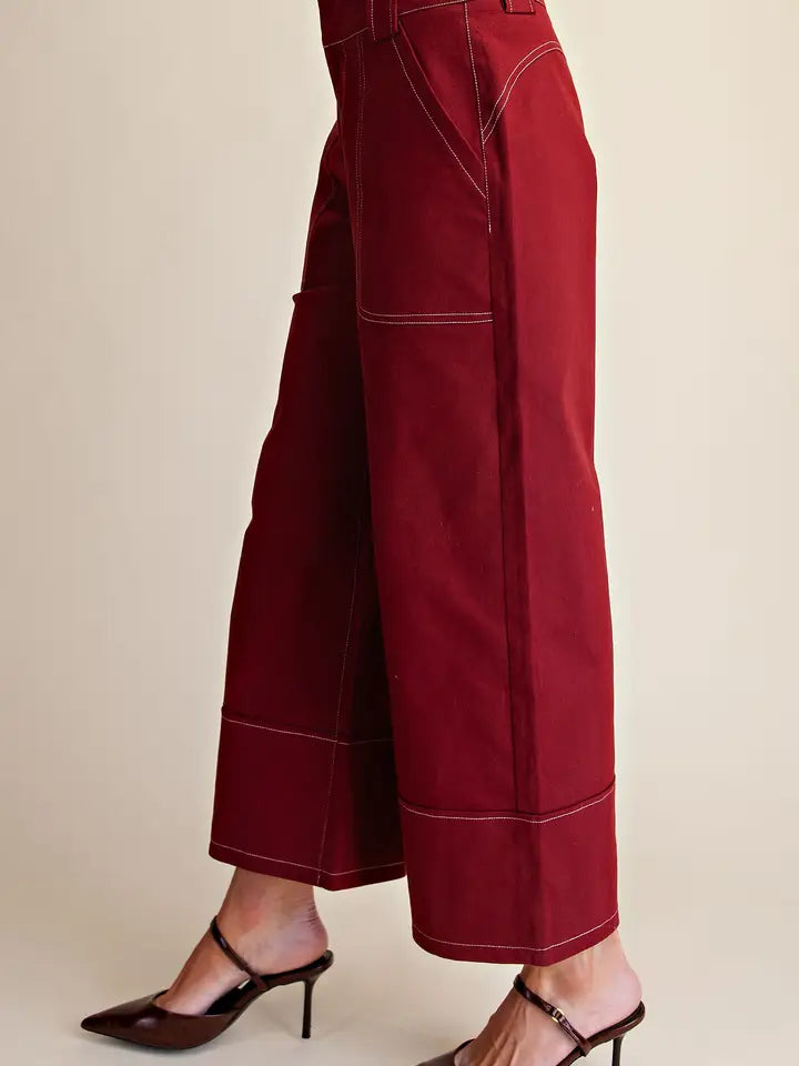 A close-up of a person wearing burgundy wide-leg cropped pants with contrast light stitching and dark brown slingback mule heels, shown against a plain beige studio background.