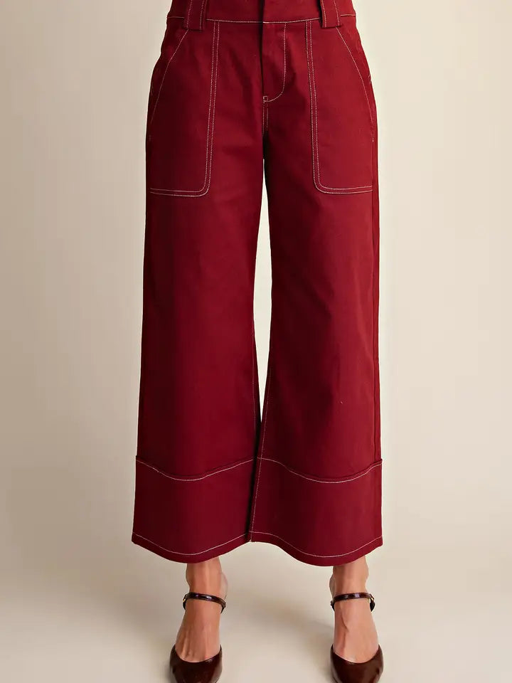 A person wearing high-waisted, wide-leg cropped pants in deep burgundy with white contrast stitching, paired with dark brown ankle-strap heels, against a plain beige studio background.