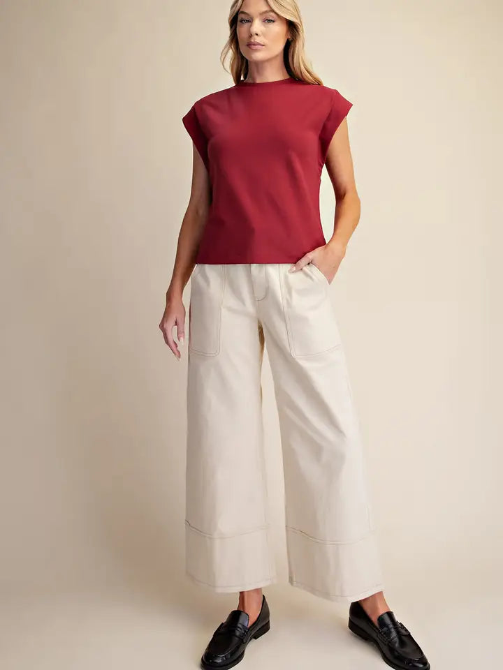 A woman modeling a sleeveless deep red top and off-white wide-leg cropped pants with black loafers, standing against a plain beige studio background.