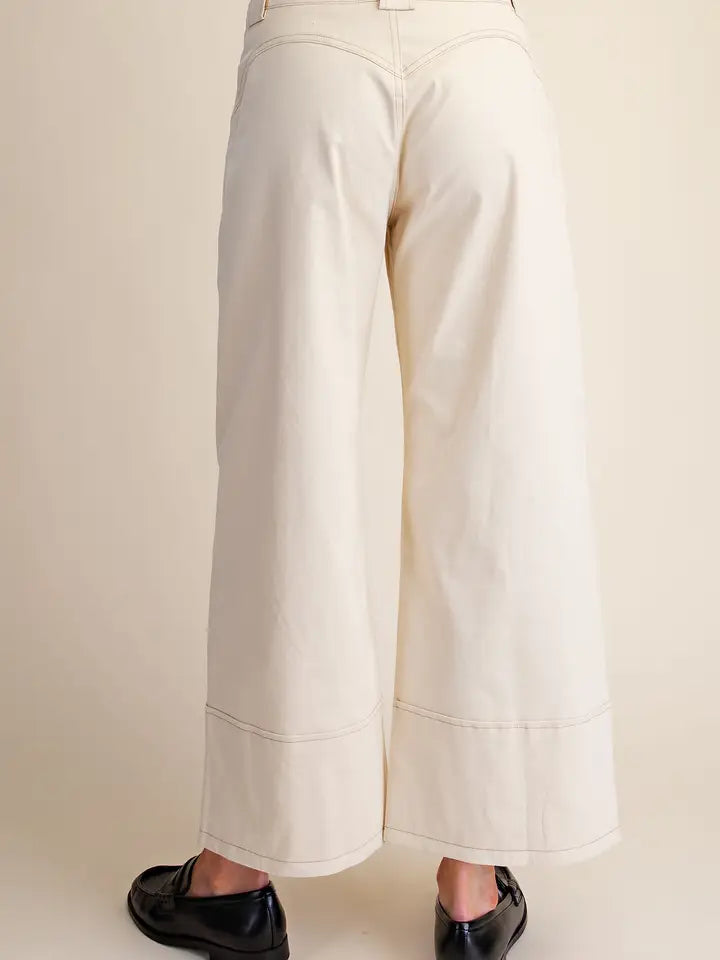 A person shown from the back wearing off-white wide-leg cropped pants with double hem stitching and black loafers, against a plain beige studio background.