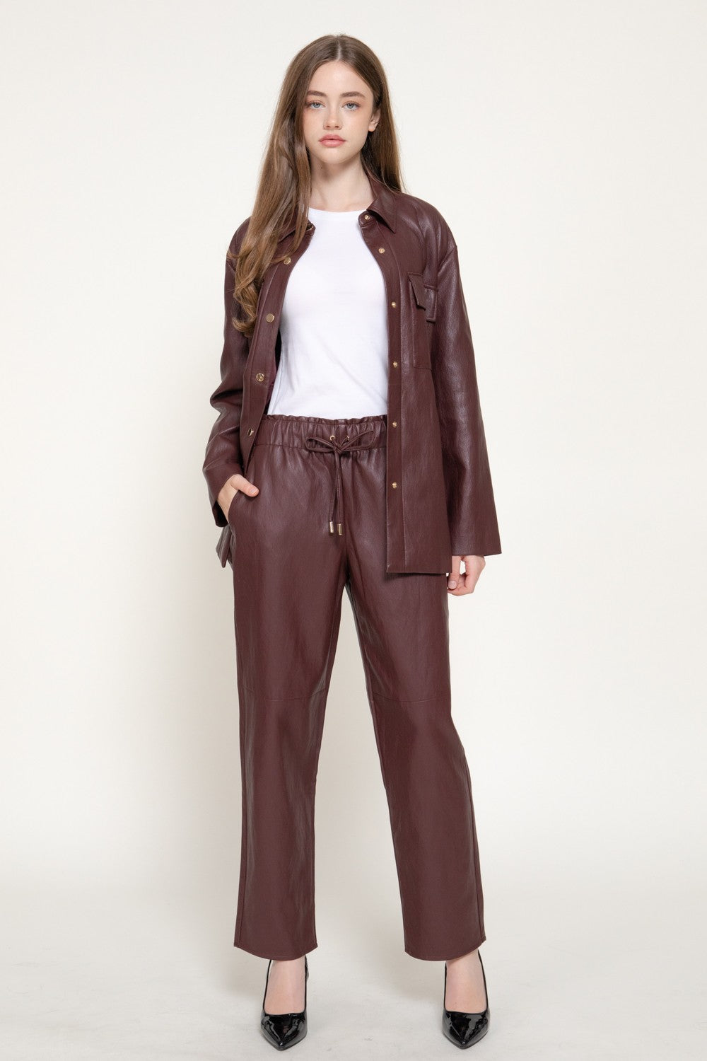 A woman modeling a matching brown faux leather jacket and wide-leg pants set, styled with a white t-shirt and black heels, against a plain off-white studio background.