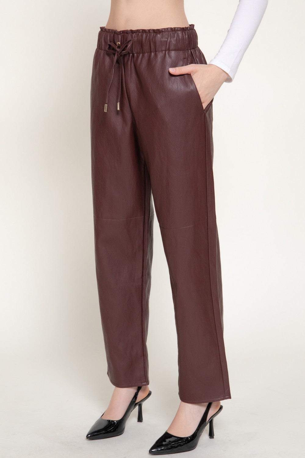 A person wearing dark brown faux-leather drawstring pants with side pockets, paired with black pointed-toe heels, shown against a plain light beige studio background.