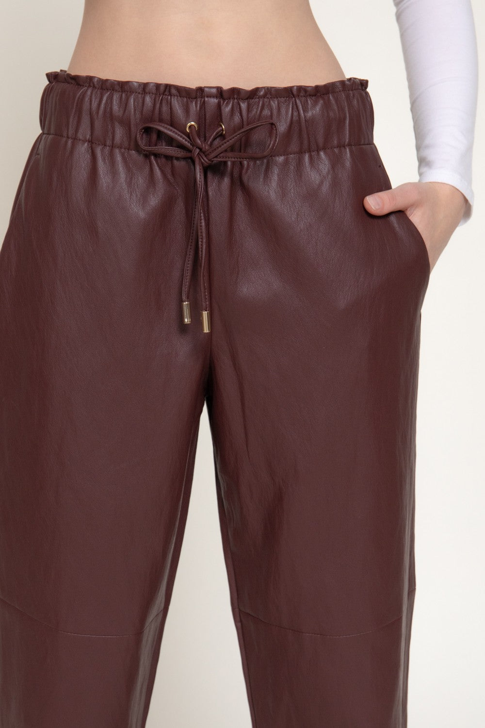 A close-up of a person wearing dark brown faux-leather drawstring pants, showing the waistband, side pocket with a hand in the pocket, and the lower leg area against a plain light background.