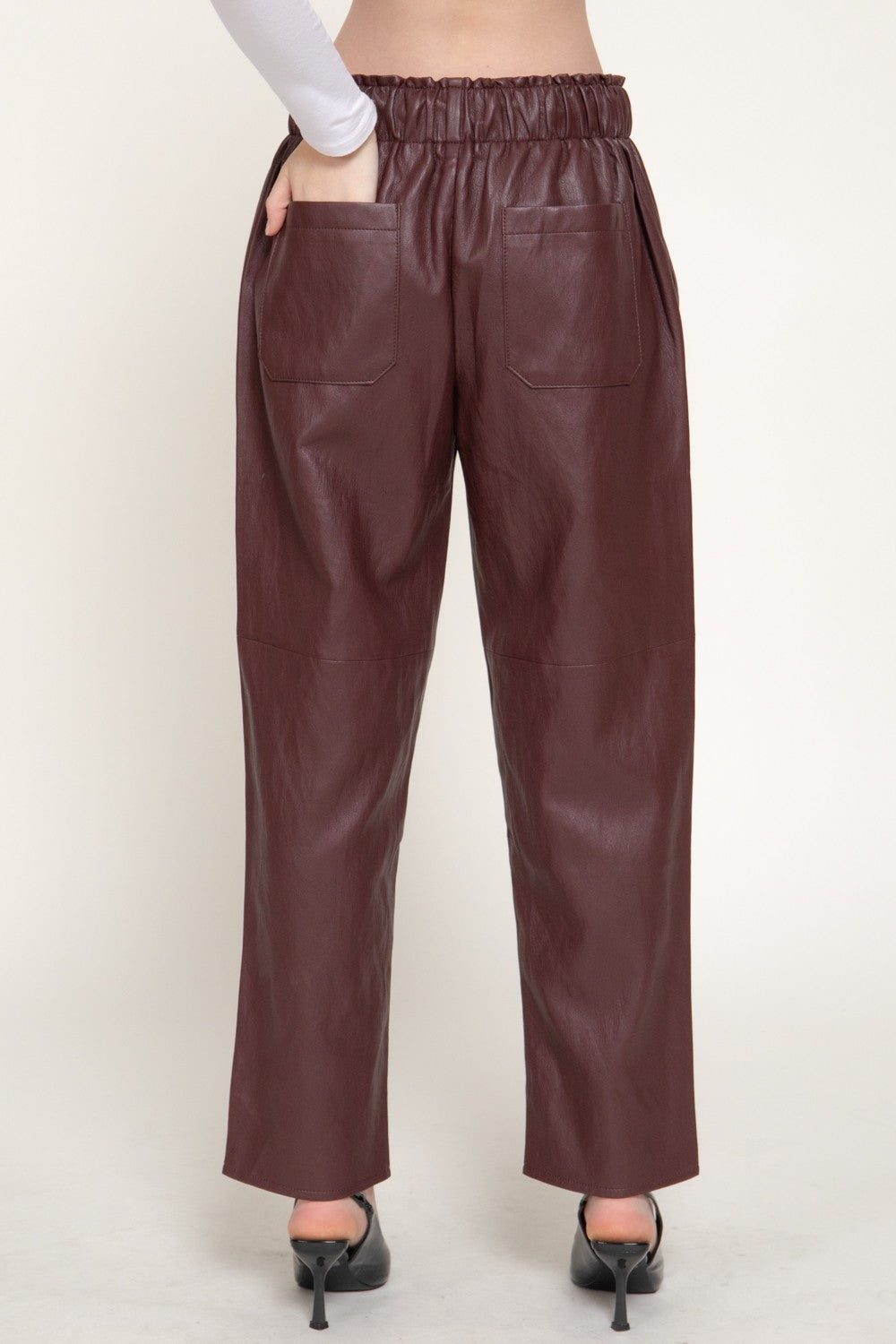 A person shown from the waist down wearing wide-leg brown faux-leather pants with an elastic waistband, side pockets, and a straight cropped leg, paired with black pointed-toe heels against a light beige studio background.