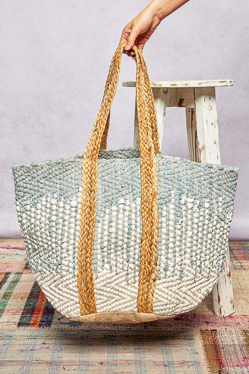 Woven beach bag with braided handles held by a hand on a colorful rug.