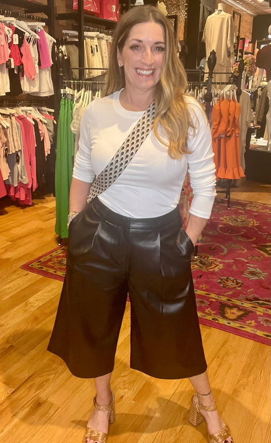 A person standing in a retail environment wearing black wide-leg cropped pants with a vegan leather finish, paired with a white top and accessorized with a handbag and a ring.