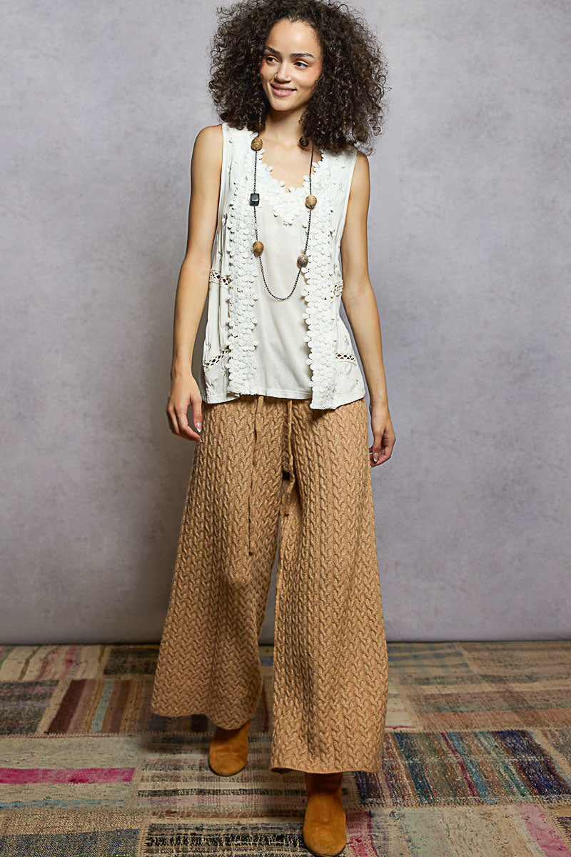 Woman wearing a sleeveless top and patterned pants standing against a gray wall.