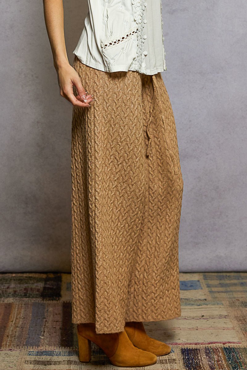 Person wearing a long brown knitted skirt with a white top against a gray wall.