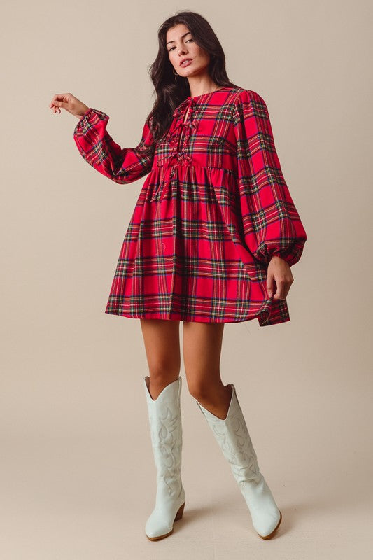 A woman wearing a red and black plaid babydoll dress with puff sleeves, a crew neckline, and front tie closures. She is also wearing white boots.