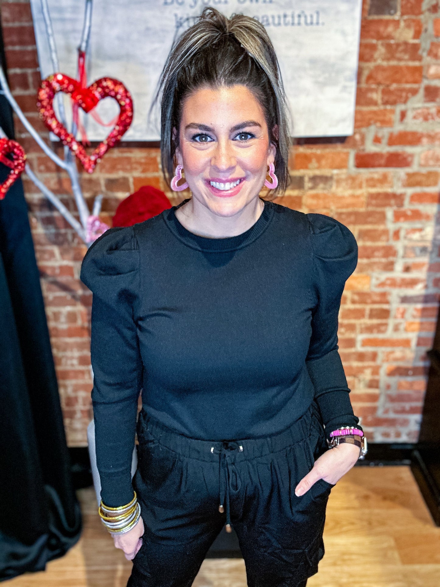 A woman wearing a black puff sleeve sweater top with black pants, accessorized with multiple bracelets and earrings.