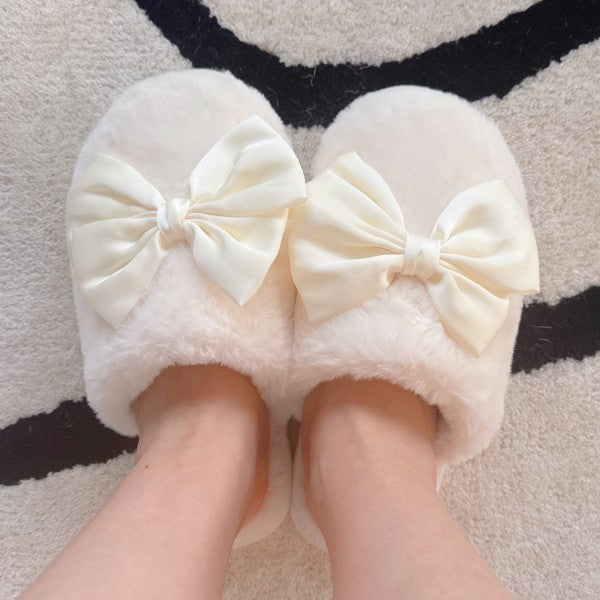 White fuzzy slippers with large cream-colored bows worn indoors on a patterned rug.