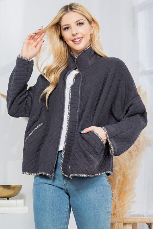 Woman wearing a dark gray quilted jacket with a white shirt and blue jeans indoors.