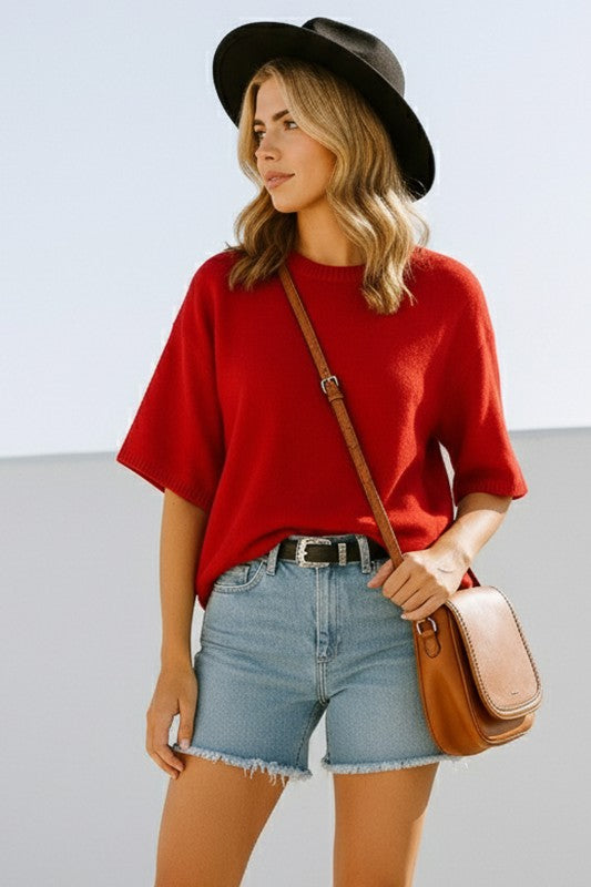 Woman wearing a red shirt, denim shorts, and a black hat with a brown leather bag.