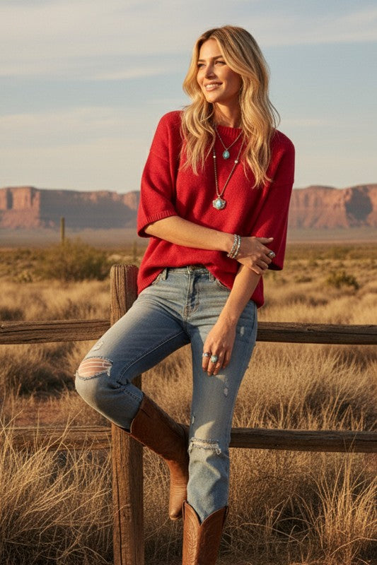Woman in a red sweater and jeans standing in a desert landscape with mountains in the background.