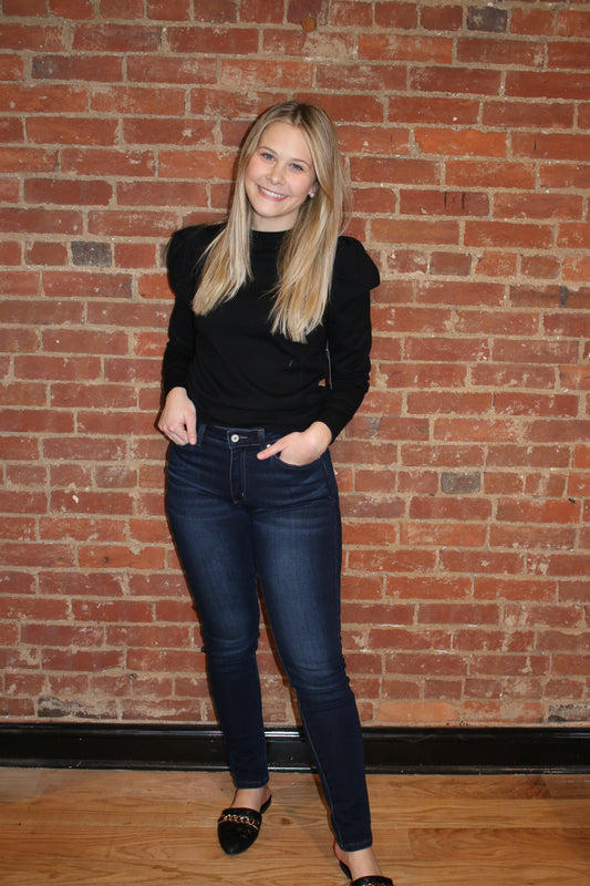 A person standing against a brick wall background, wearing dark wash skinny jeans and a black top.