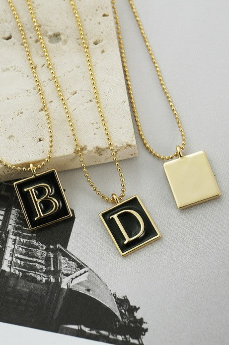A gold ball chain necklace with a square black enamel charm featuring a gold letter accent, displayed against a neutral background.